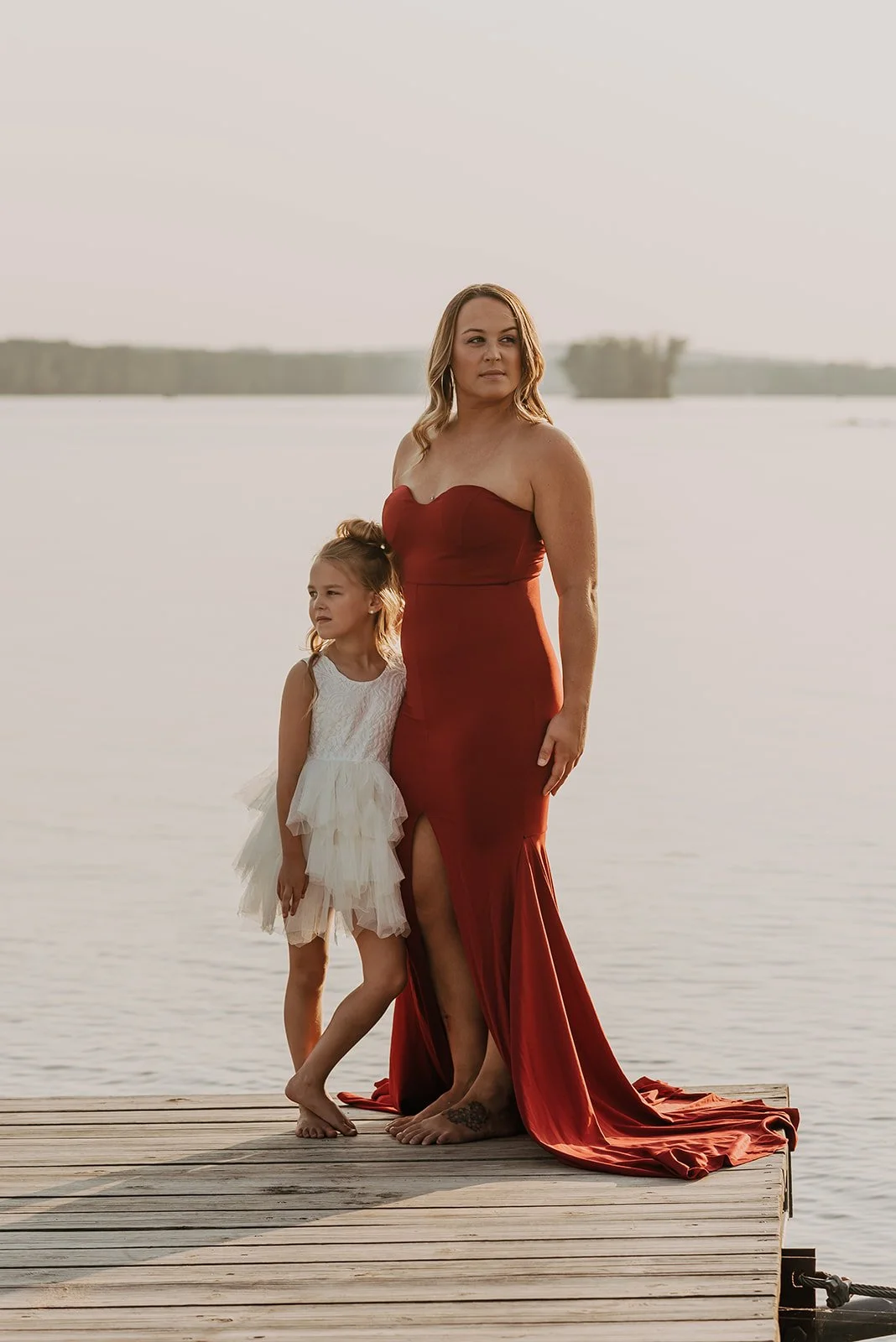 A woman in a red evening gown and a young girl in a white dress stand on a wooden dock by a body of water at sunset.