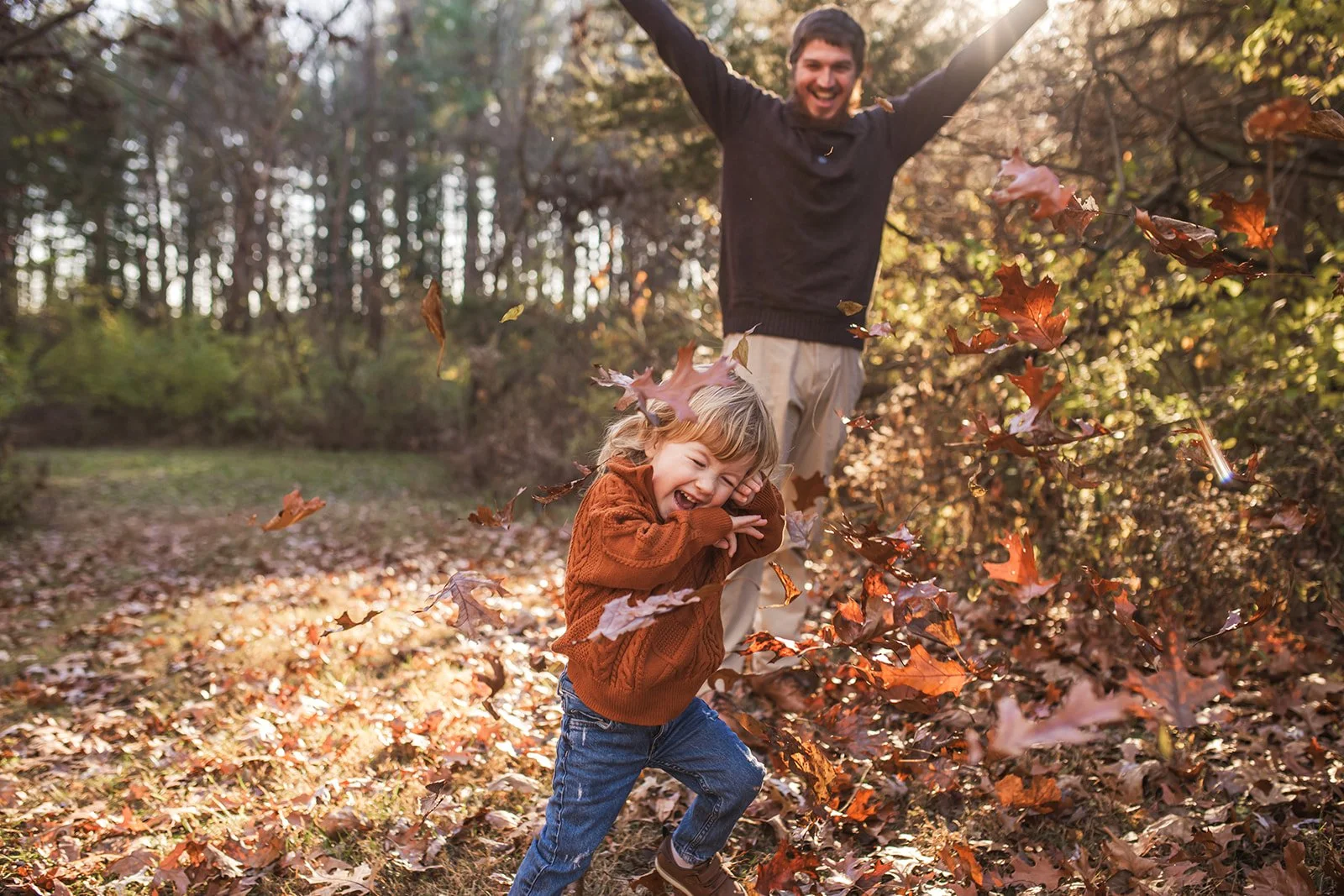 A man and a young girl playing with falling leaves in a wooded area during autumn, with the girl joyfully crouching and the man enjoying in the background.