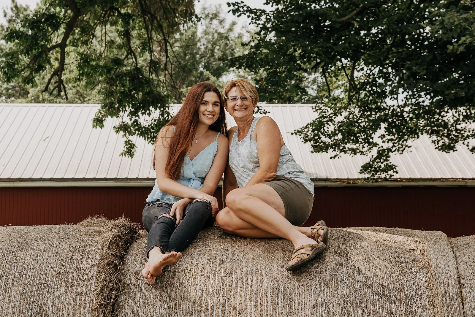 A young woman with long red hair and an older woman with short blonde hair sitting together on a hay bale outdoors, smiling at the camera. The young woman wears a light blue top and ripped jeans, and the older woman wears a sleeveless top and shorts.