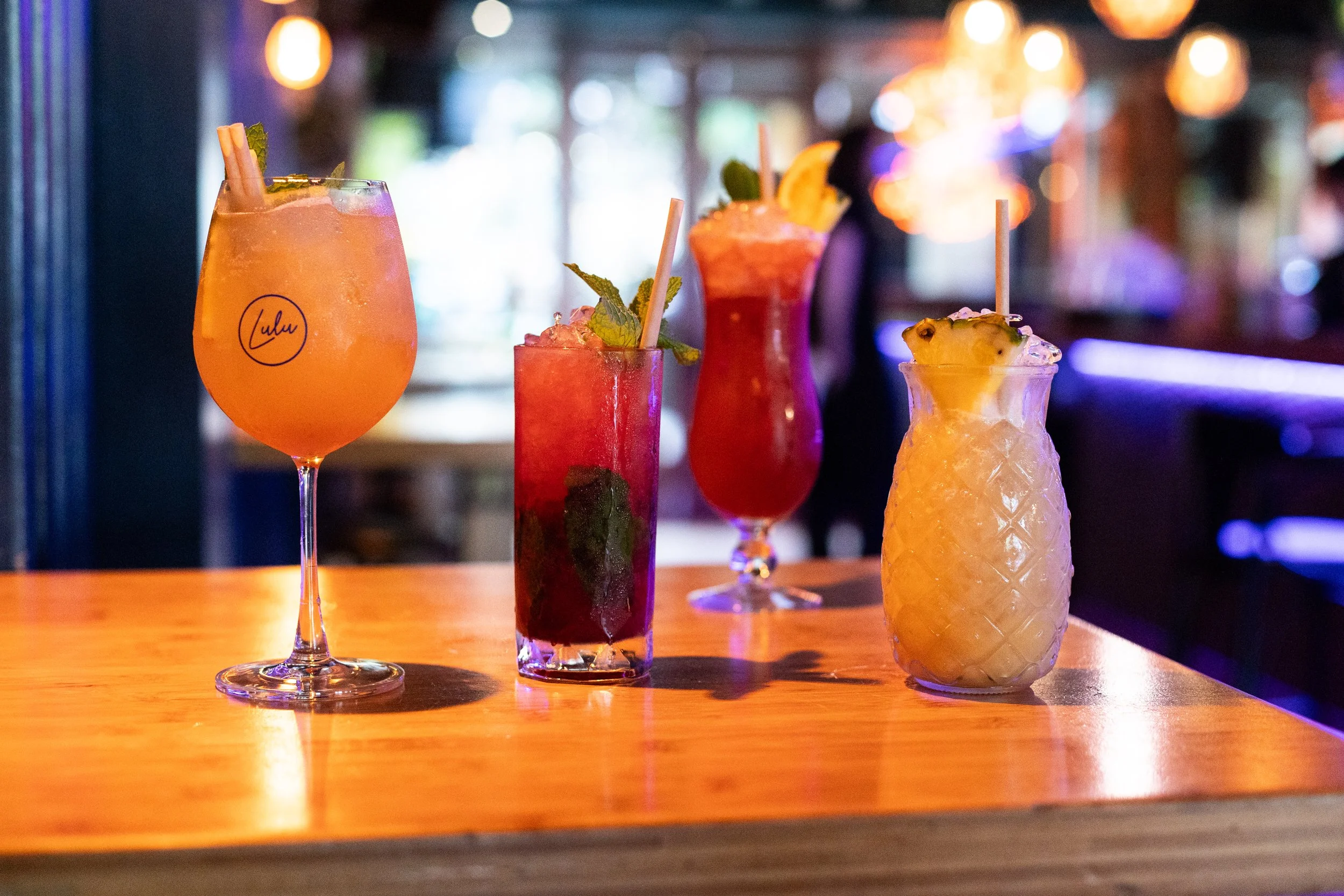 An assortment of colorful cocktails on a wooden bar in a dimly lit bar or restaurant with blurred background and warm lighting.
