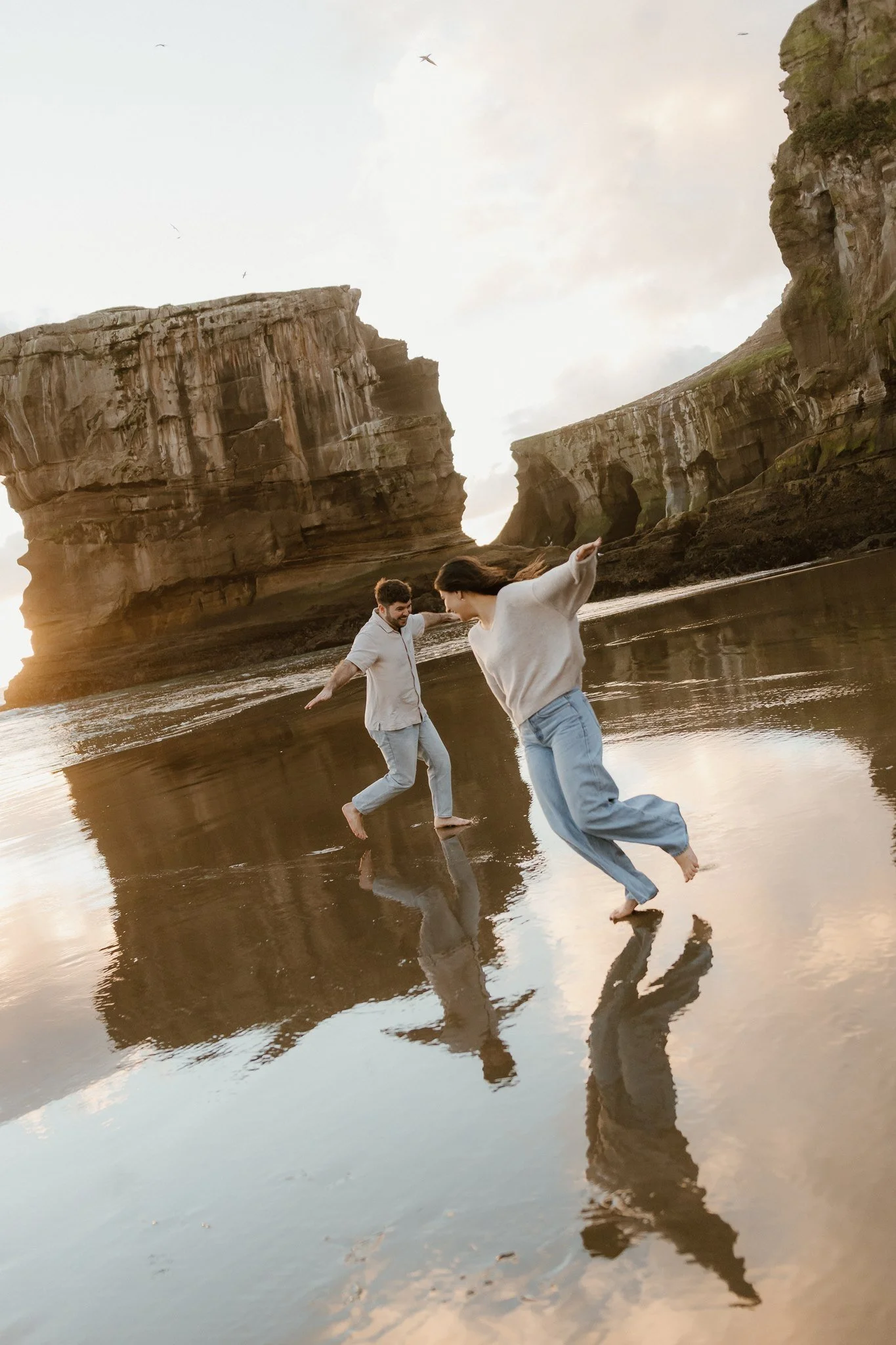 A couple playing and holding hands on the wet sand of a beach during sunset, with large cliff formations in the background and reflections of their figures on the water.
