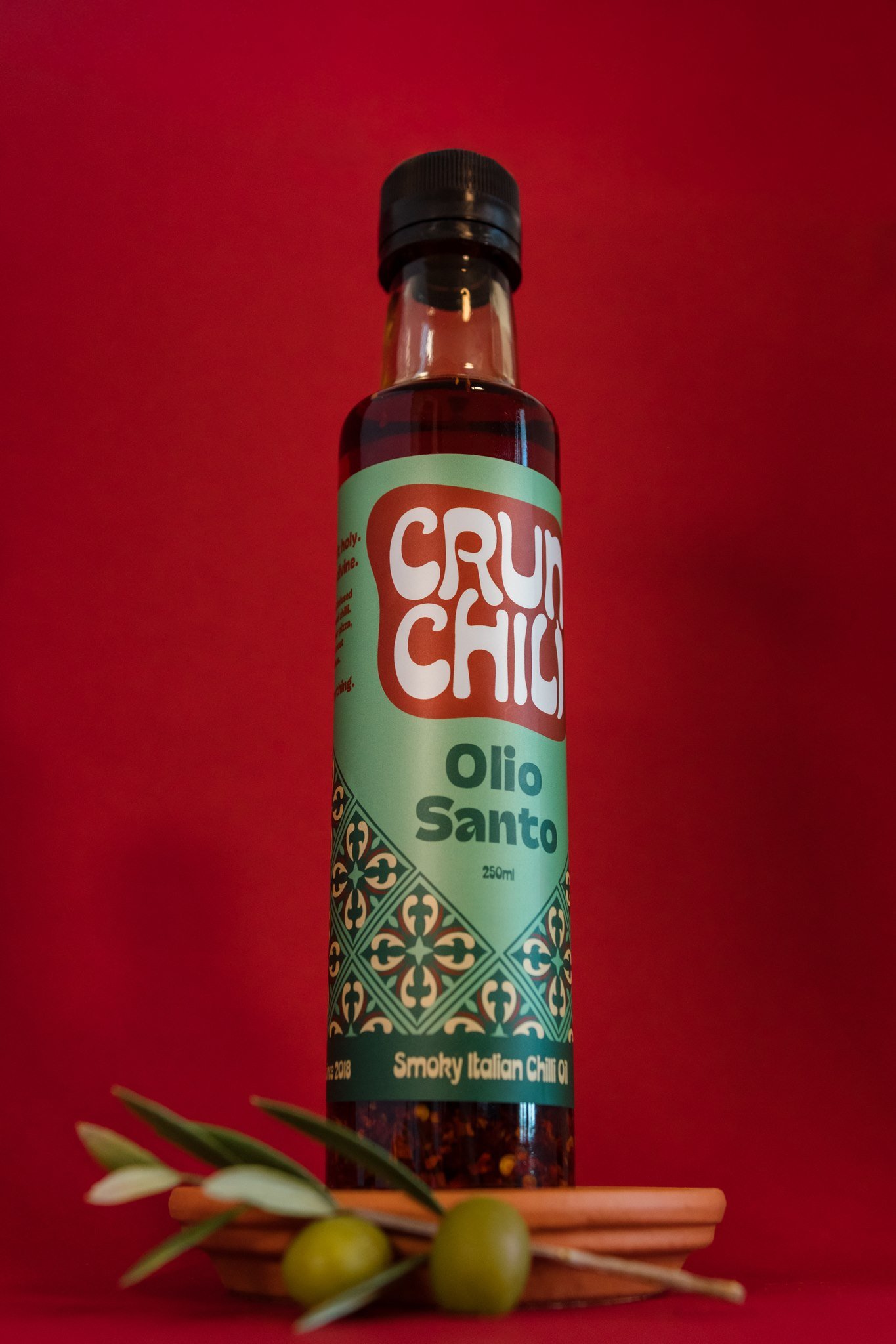 A bottle of Cru Shok Olio Santo seasoning with a red background, placed on a small round wooden tray. There are some green olives with leaves at the bottom of the tray.