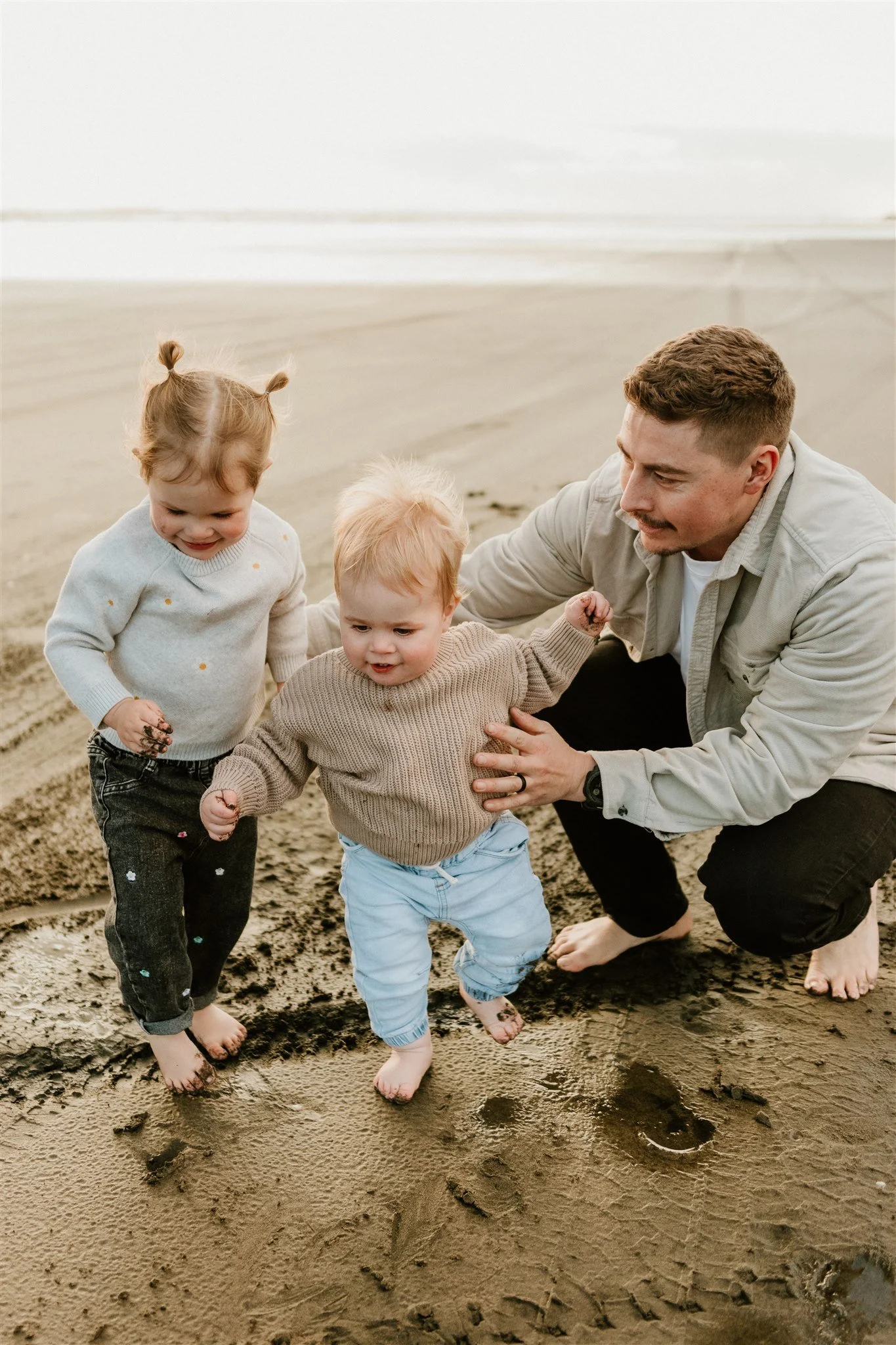 A man and two small children barefoot on a sandy beach, playing and looking at footprints and holes in the sand.
