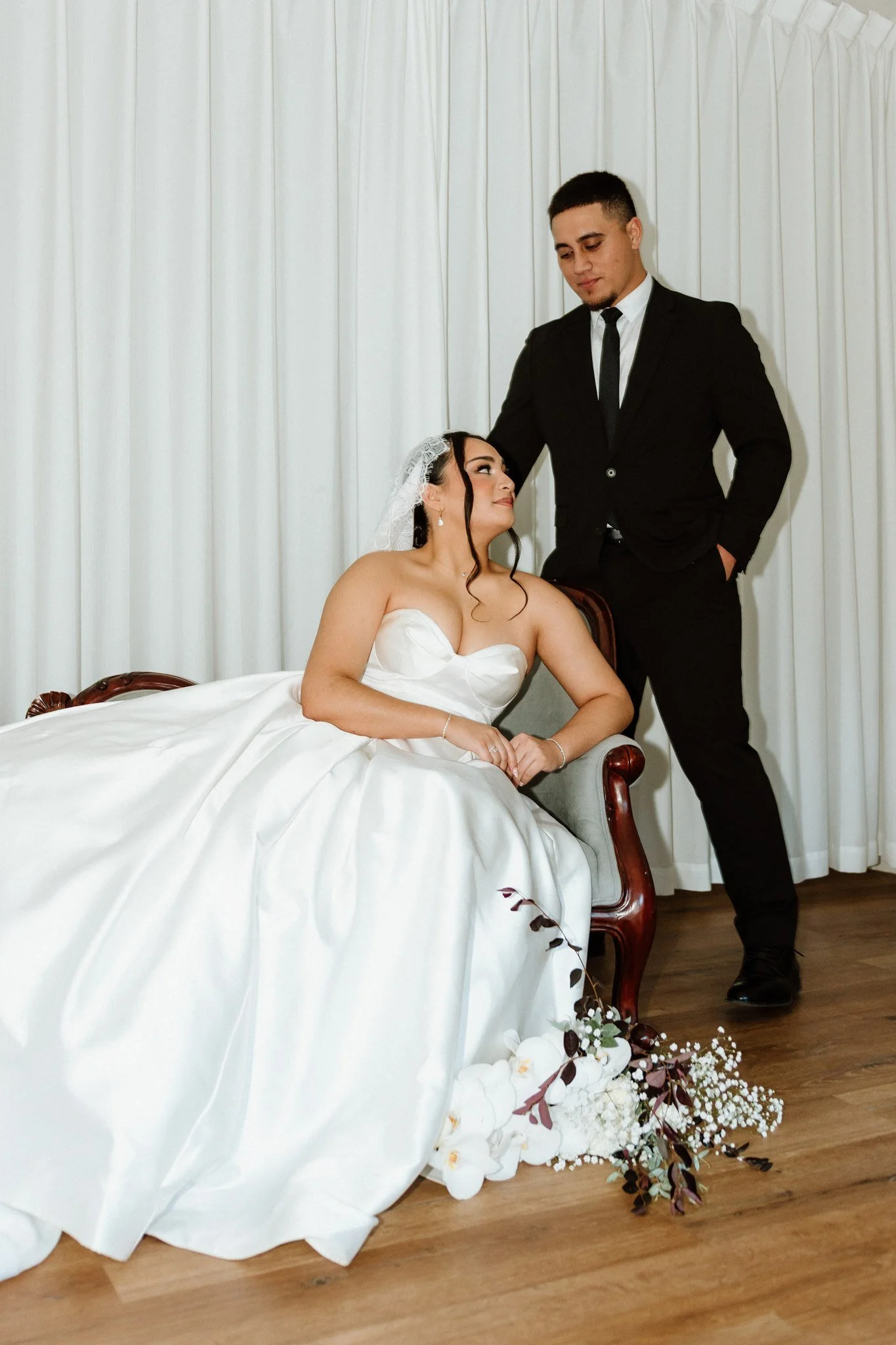 A bride in a white wedding dress sitting on a sofa, and a groom in a black suit standing next to her, resting his hand on her head, both looking at each other. There is a bouquet of flowers on the floor in front of them.