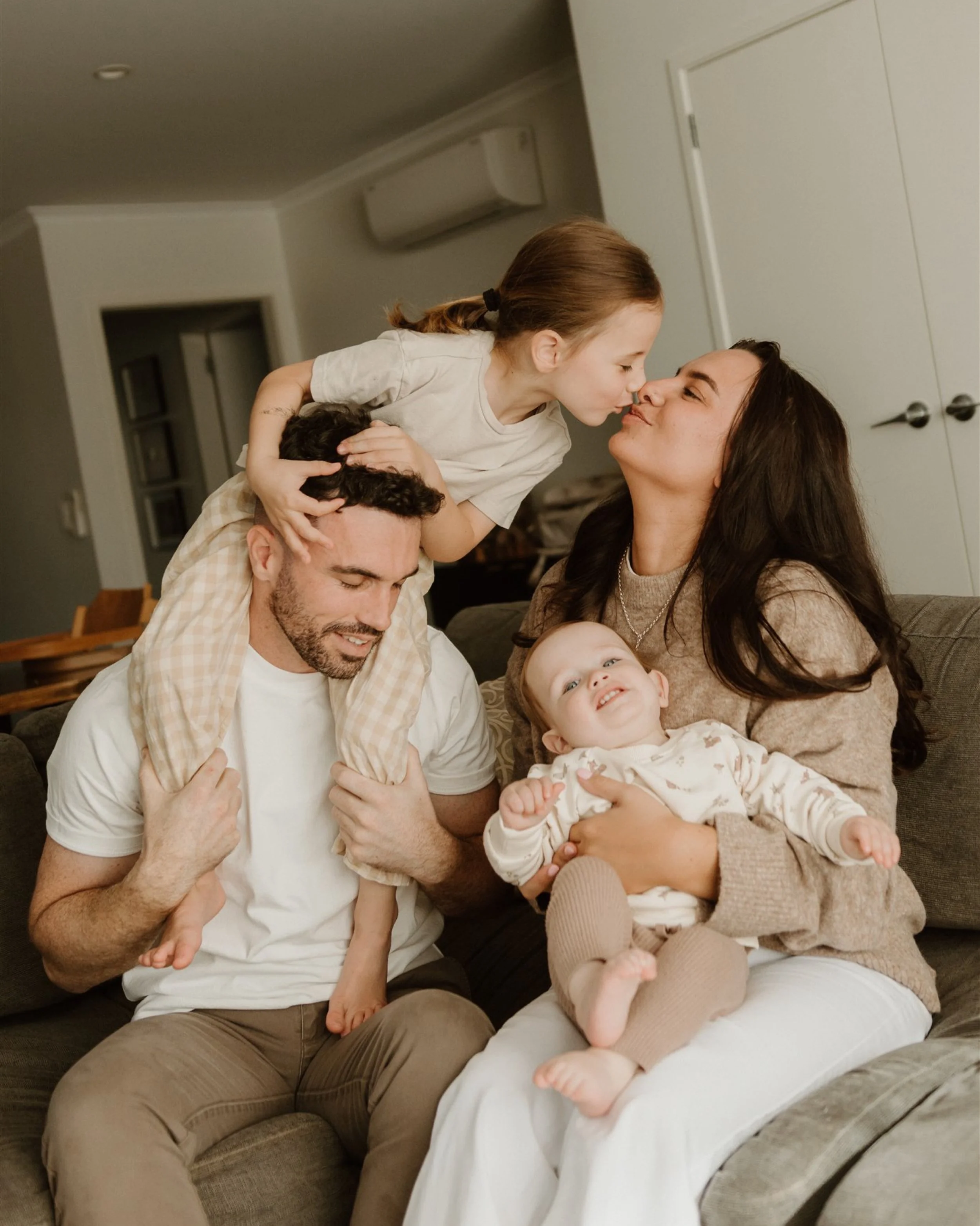 Family of four with two young children sitting together in a living room, sharing a happy moment with the children kissing and smiling.