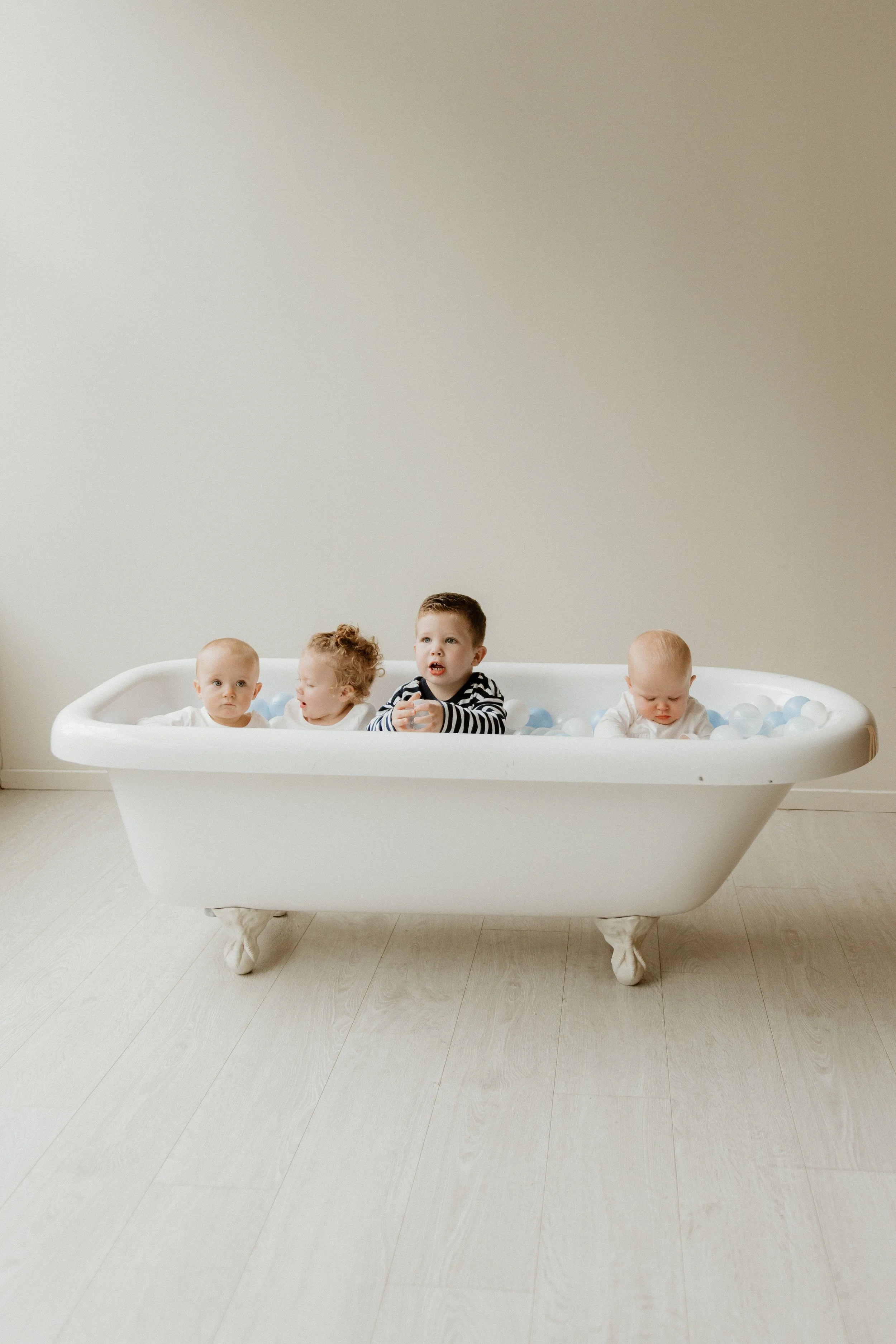 Five young children sitting in a white clawfoot bathtub filled with light blue and white plastic balls, in a room with beige walls and light wood floor.