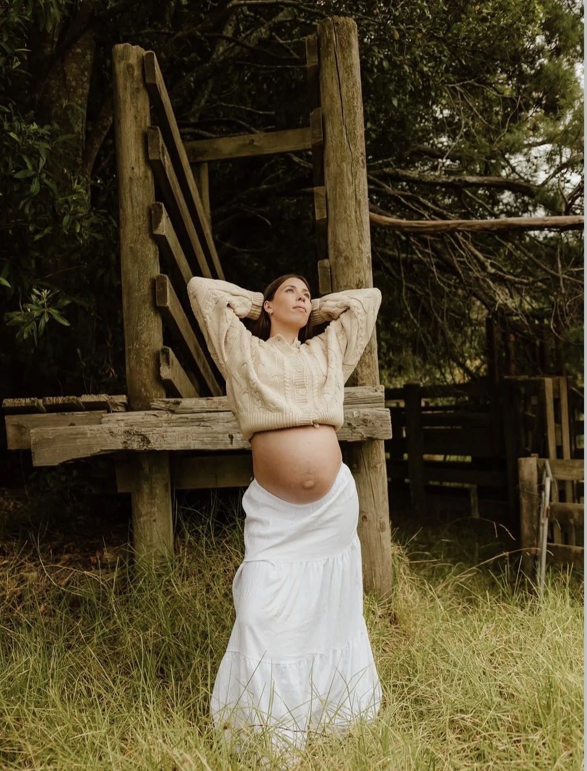 A pregnant woman standing outdoors in front of a wooden structure, wearing a beige sweater and a white skirt, with her hands behind her head, looking thoughtfully up.