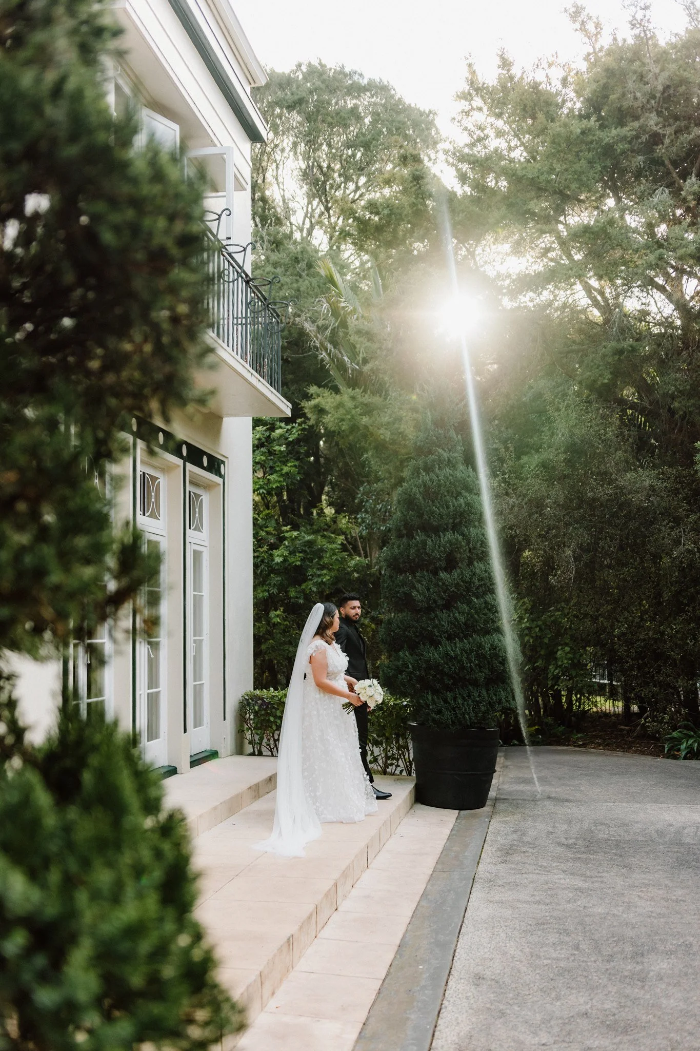 A bride and groom stand together outdoors on wedding day, with sunlight shining through trees behind them, near a white building with a balcony.