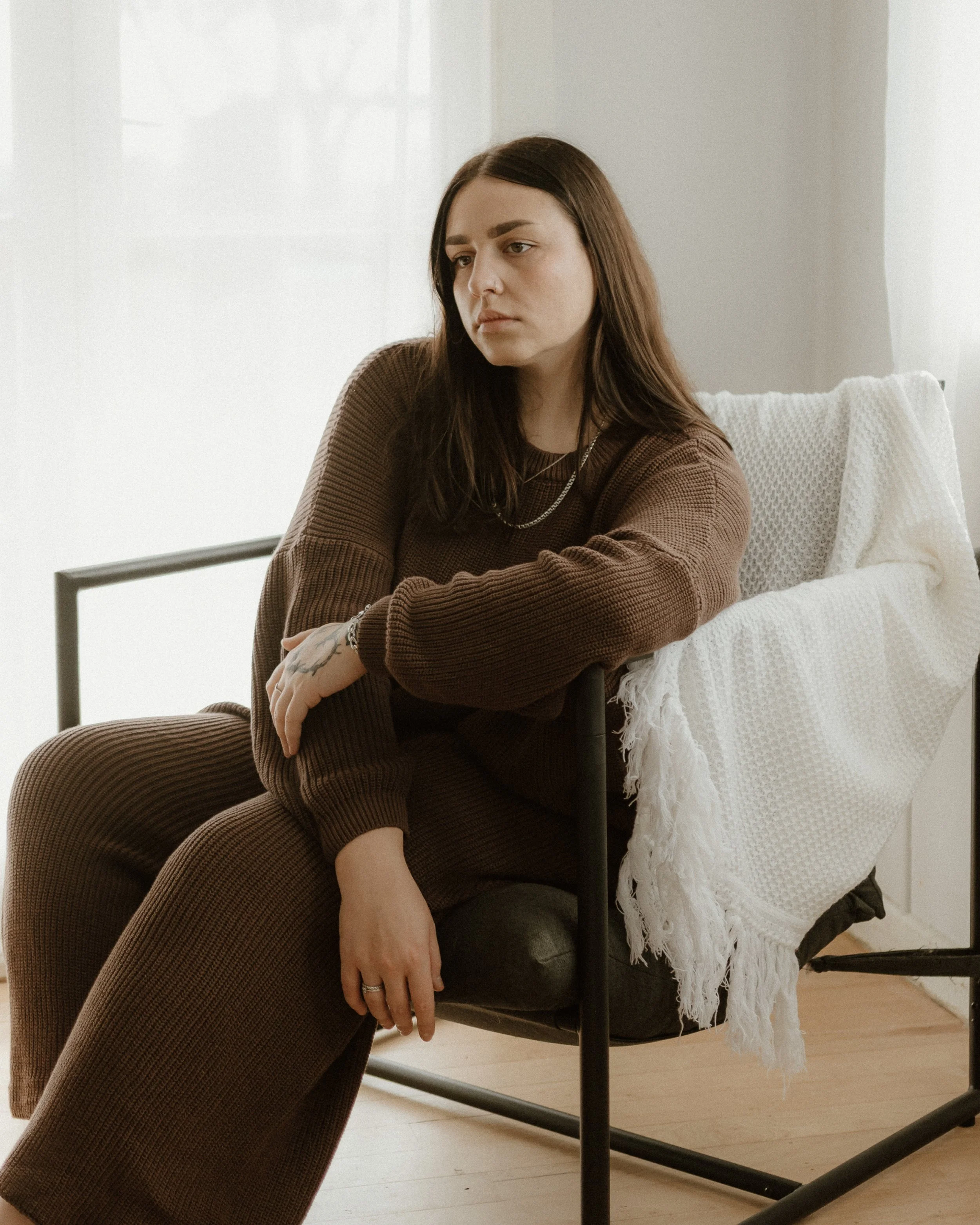 A woman with long brown hair and tattoos on her left hand sitting on a chair, wearing a brown sweater and jewelry, with a white textured blanket draped over the back of the chair, in a bright room with white walls and wooden flooring.