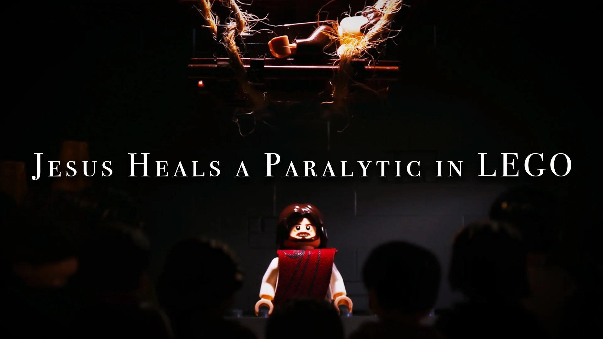 A scene from a play or event featuring a LEGO figure of Jesus and the paralytic being brought down from the roof. The scene includes the audience and dark surroundings.