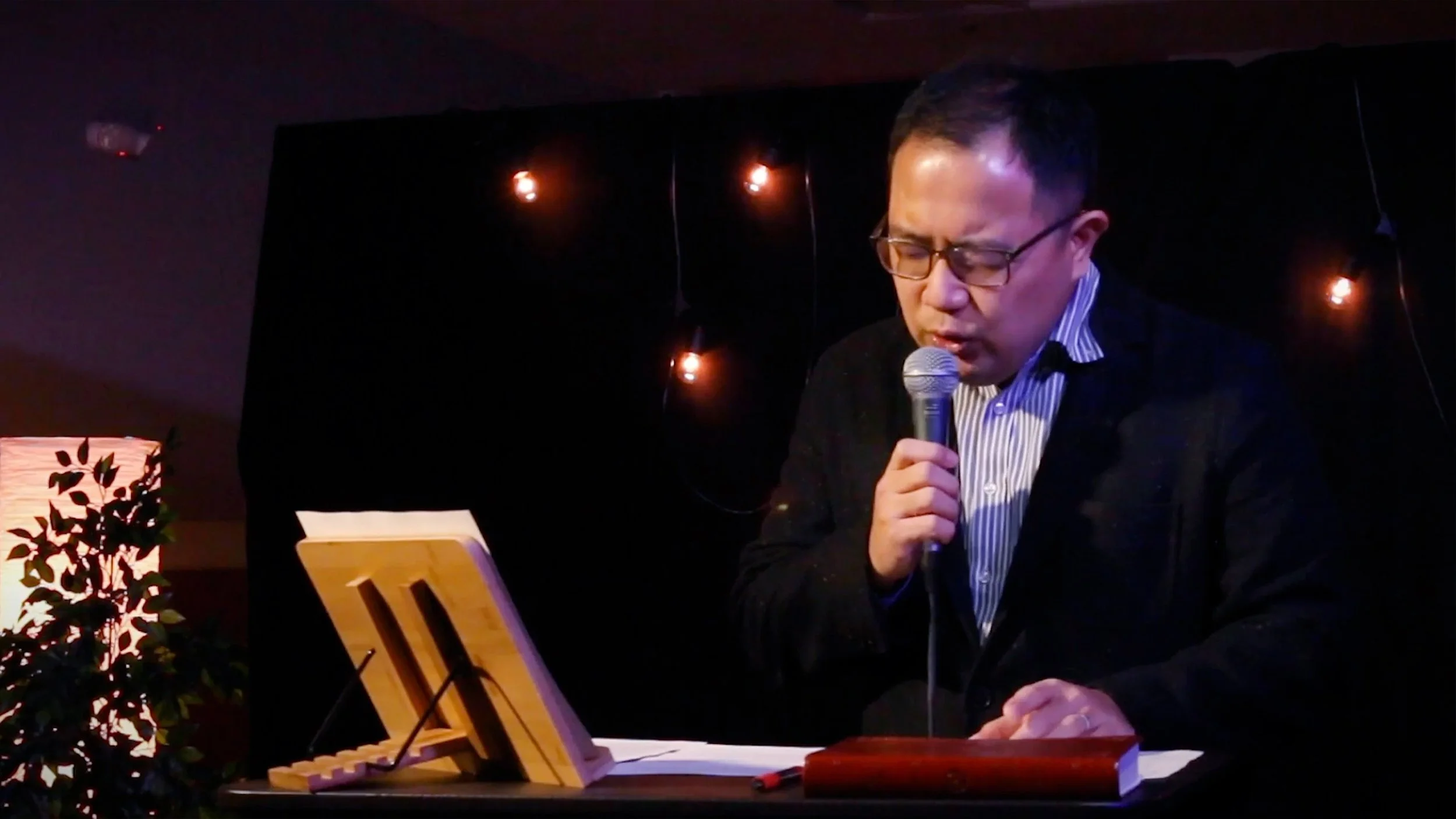 A man with glasses and short dark hair speaking into a microphone in a dimly lit room decorated with string lights, with a music stand and a small potted plant on the table in front of him.