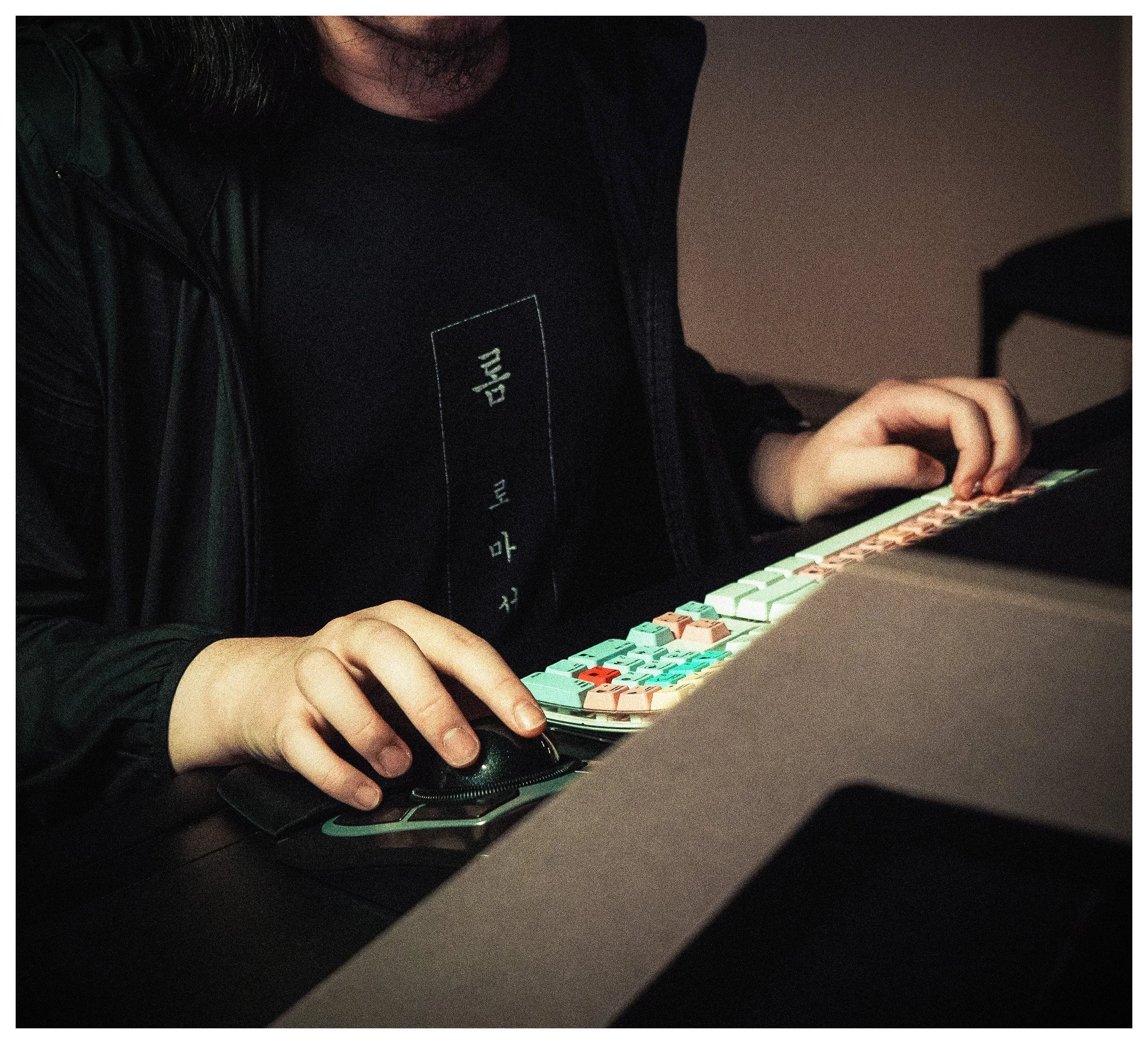 Person using a phone with colorful keyboard keys, wearing a black shirt with text and a dark jacket, in a dimly lit environment.