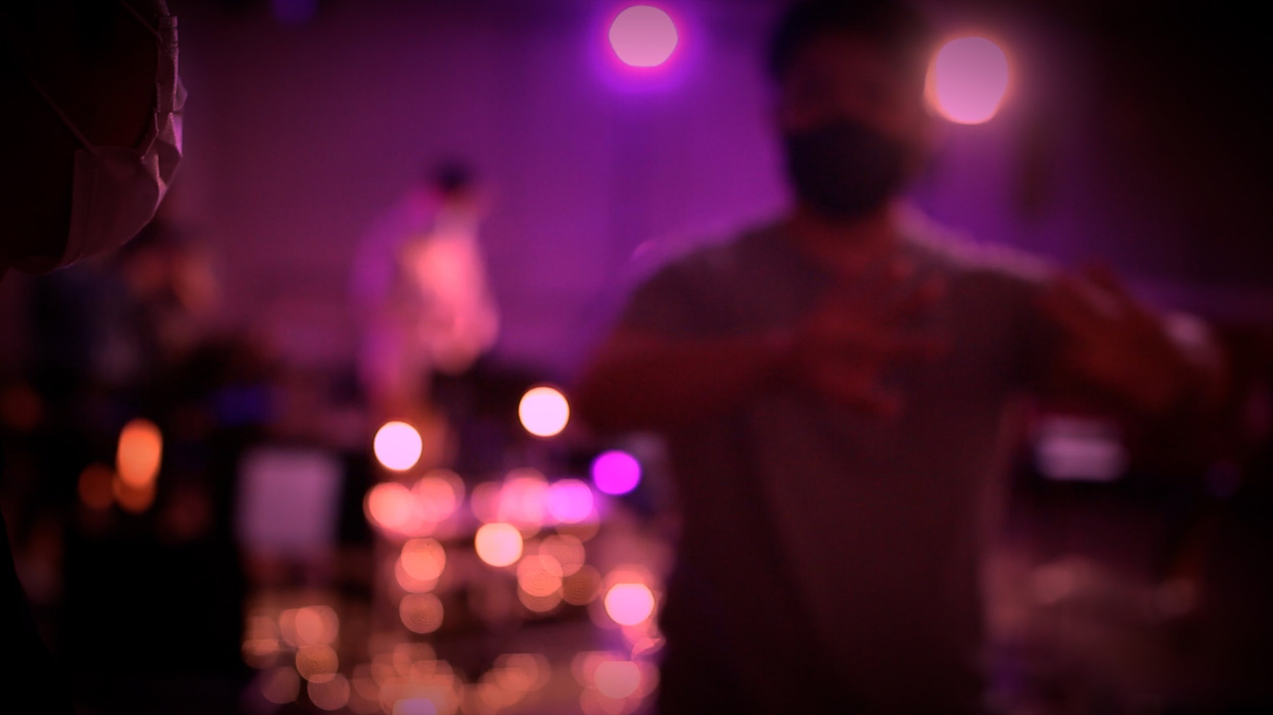 Blurred figures of people in a dimly lit environment with purple and pink lighting, creating a bokeh effect.