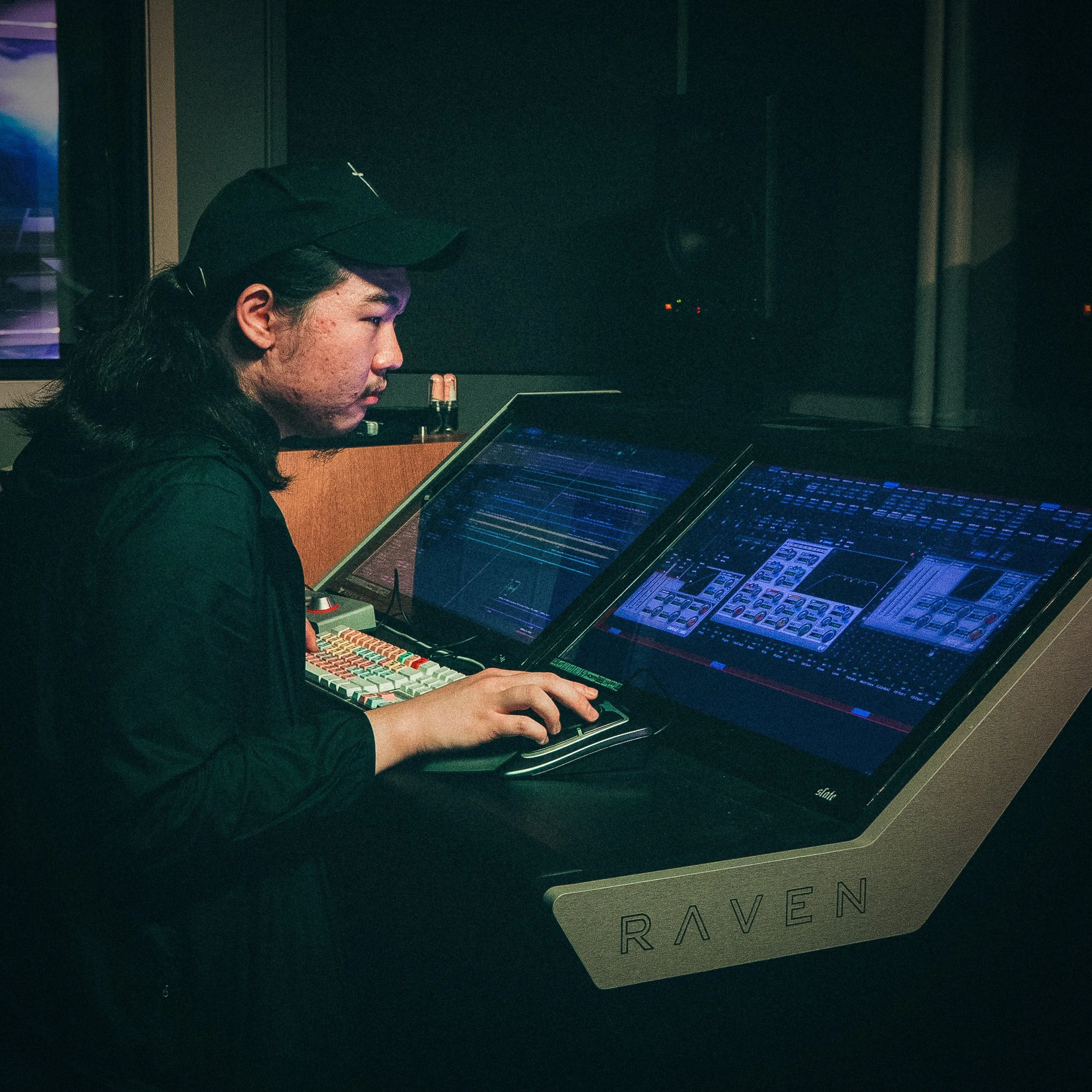 A person with long dark hair wearing a black cap is working on a music production device with a laptop, in a dark room.