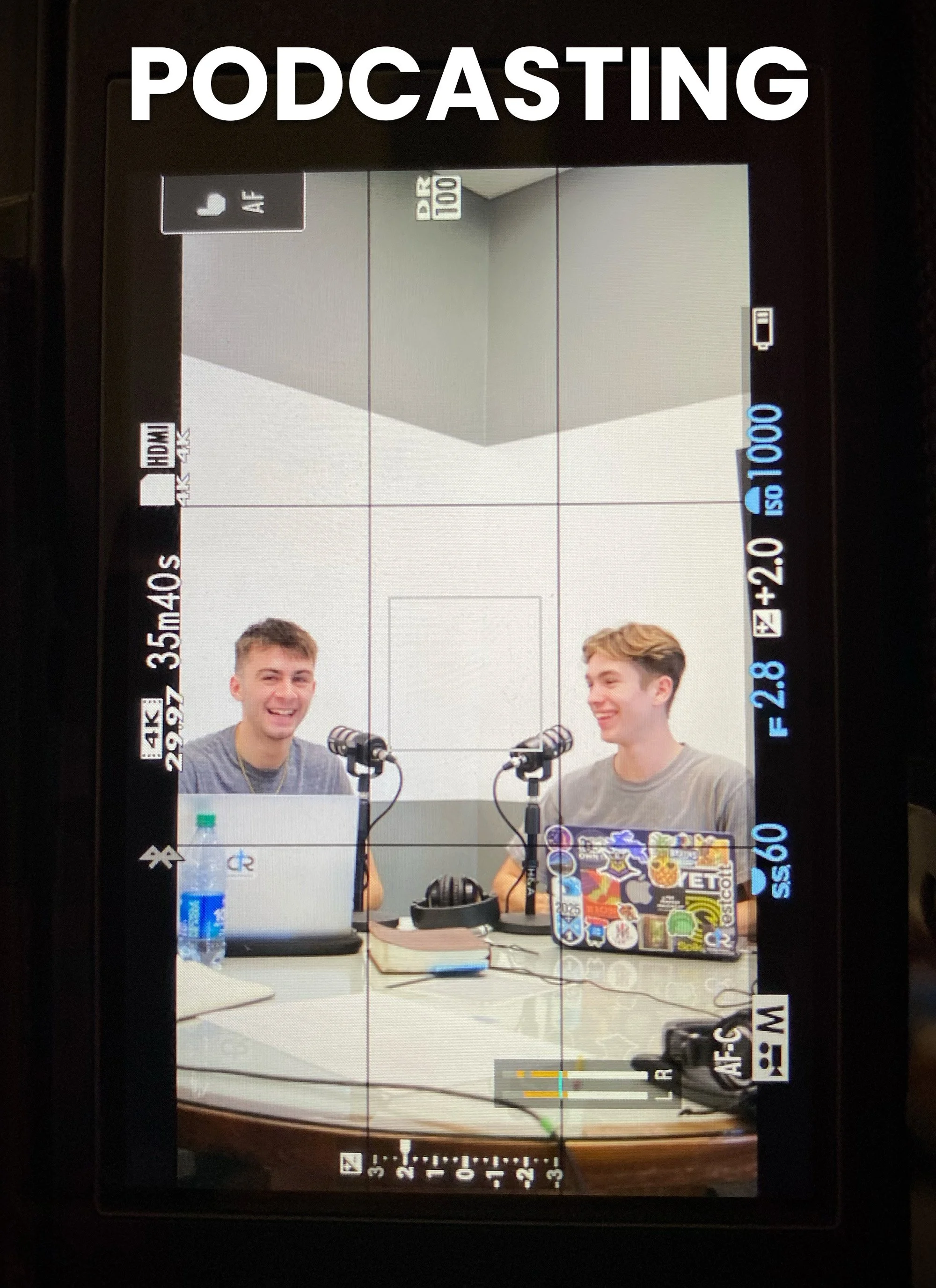 Two young men sitting at a table with microphones, laptops covered with colorful stickers, and water bottles, in a podcast recording setup.