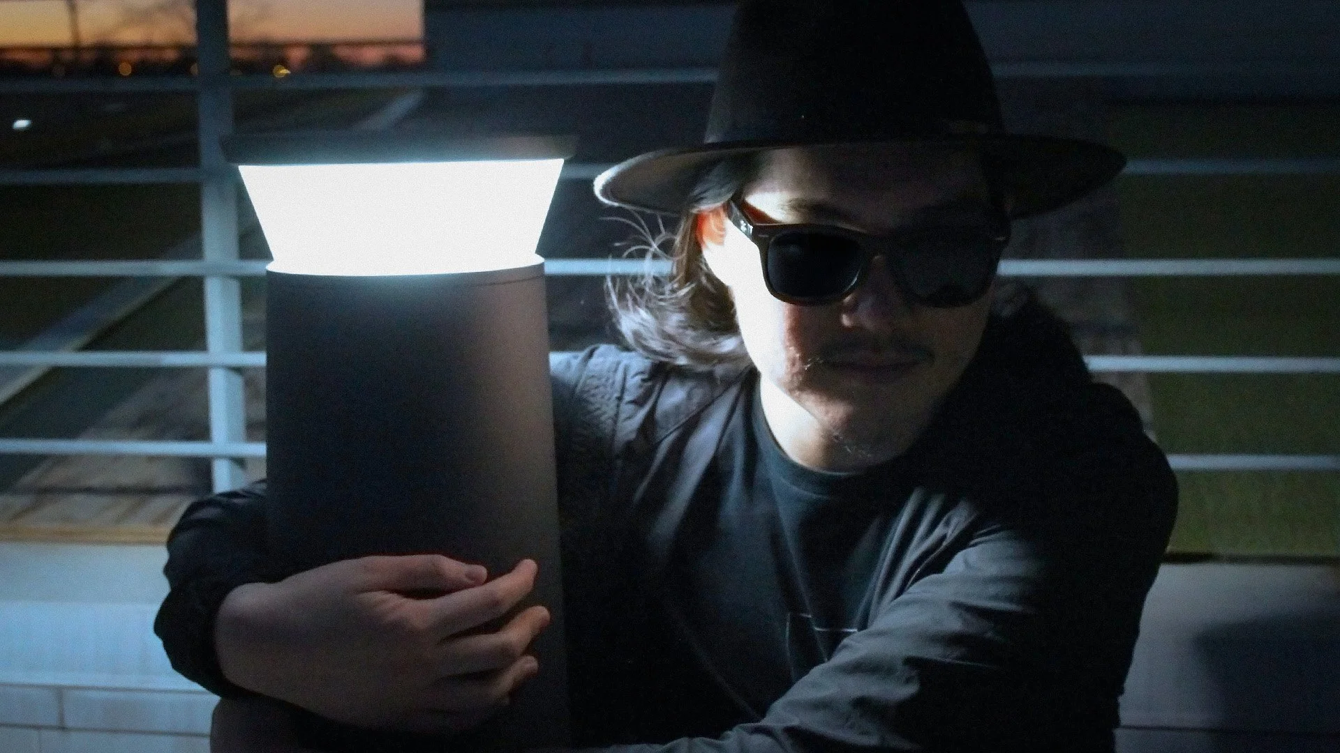 A person wearing a black hat, sunglasses, and a dark jacket holding a cylindrical object near a bright light, with a metal railing in the background.