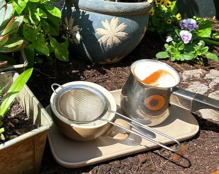 A tray with a metal coffee pitcher topped with foam and cinnamon, a metal strainer, and a beige cup. Surrounding plants and flowers in pots in a garden setting.