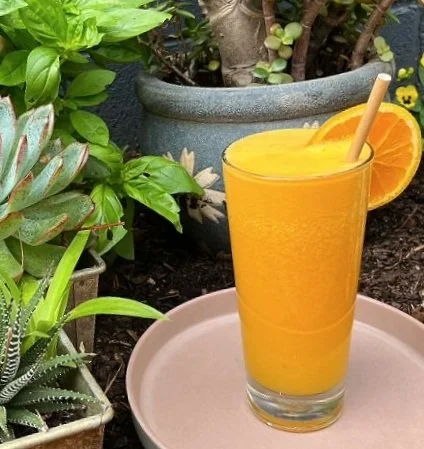 Glass of orange juice with orange slice and straw, placed on a pink tray, surrounded by potted plants.