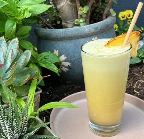 A tall glass of pineapple smoothie garnished with a pineapple wedge. The glass is placed on a pink tray with various potted plants in the background.