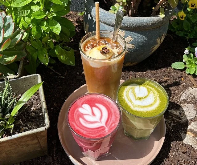 Three colorful lattes with latte art on a pink tray outdoors among potted plants.