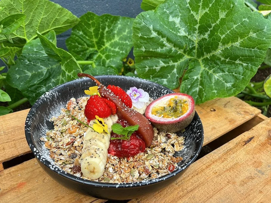 Bowl of granola topped with strawberries, banana slices, passion fruit, and edible flowers, set on a wooden surface with large green leaves in the background.