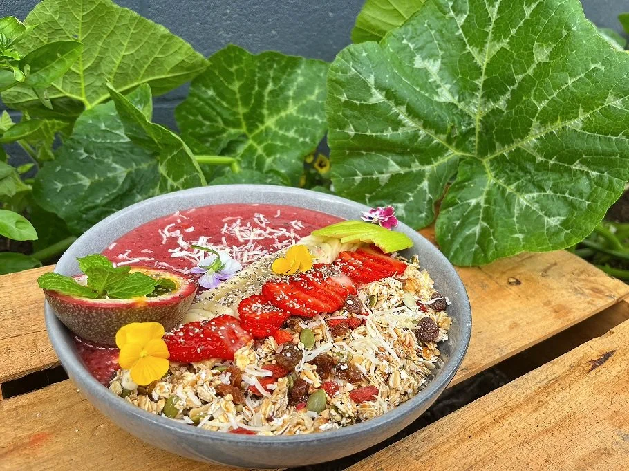 A bowl of smoothie with sliced strawberries, avocado, shredded coconut, granola, and edible flowers, with a lime half filled with a red smoothie garnished with mint leaves, placed on a wooden surface surrounded by large green leaves.