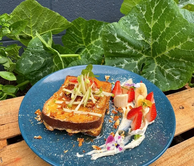 A blue plate with a slice of french toast topped with apple slices and shredded vegetables beside sliced strawberries, bananas, whipped cream, and edible flowers on a wooden surface with large green leaves in the background.