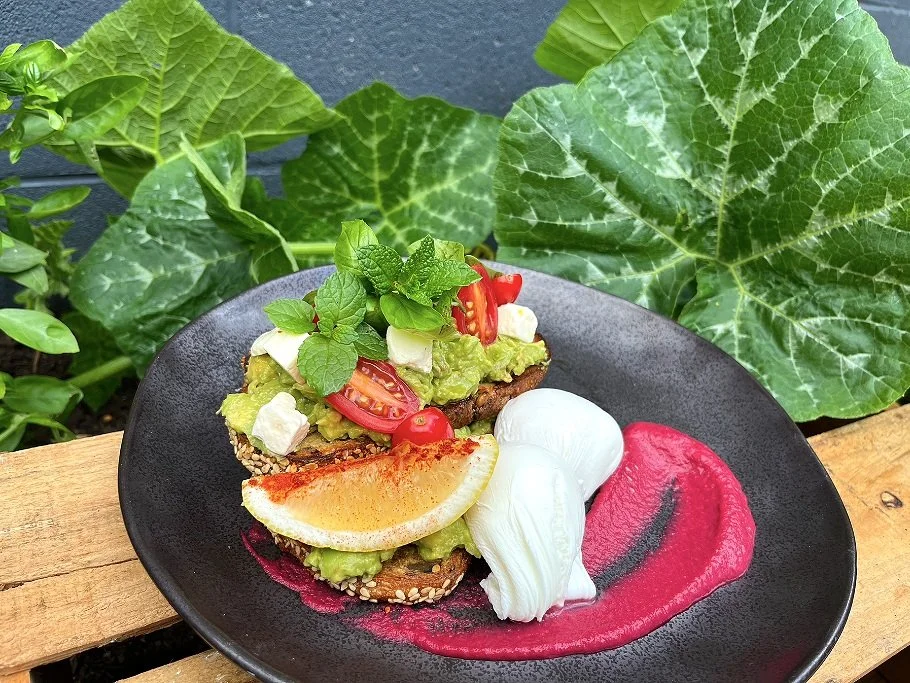 Open-faced sandwich with avocado, cherry tomatoes, cheese, mint, and a lemon wedge on a toasted bread slice. Served with dollops of white cheese, beetroot sauce, and a pink-red sauce on a black plate, with green leafy plants in the background.