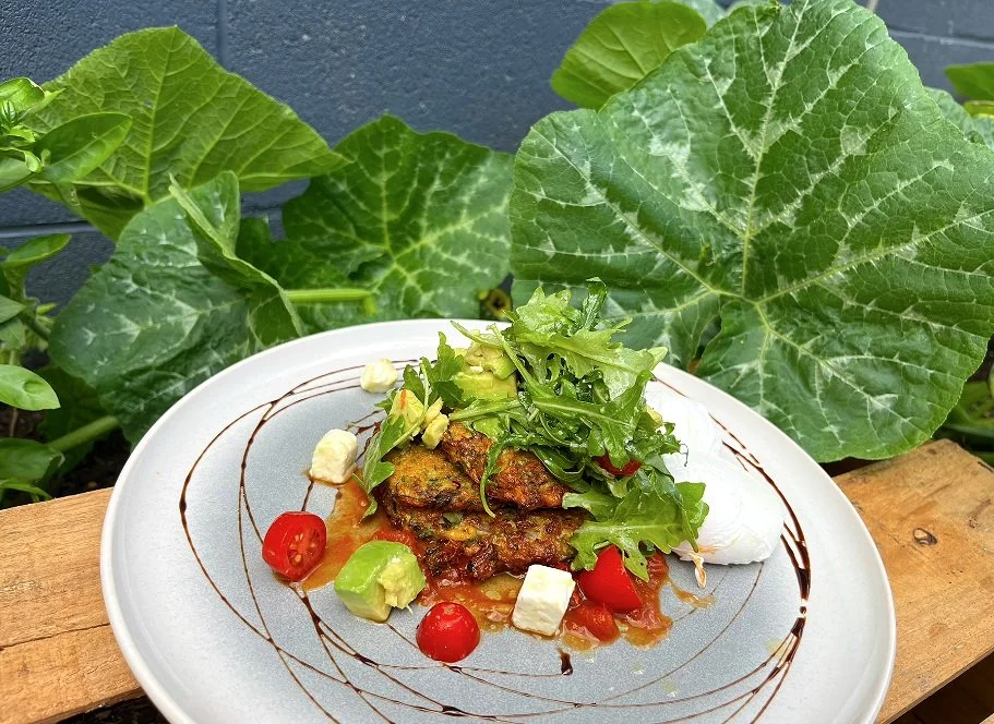 Plate of grilled veggie burger with fresh greens, cherry tomatoes, and feta cheese, garnished with balsamic glaze on a gray plate, outdoor setting with large green leaves in the background.