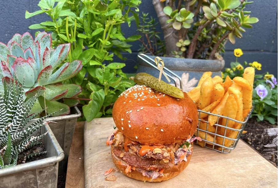 A burger with a pickle on top, served with French fries in a small metal basket, placed on a wooden board outdoors surrounded by potted plants and flowers.
