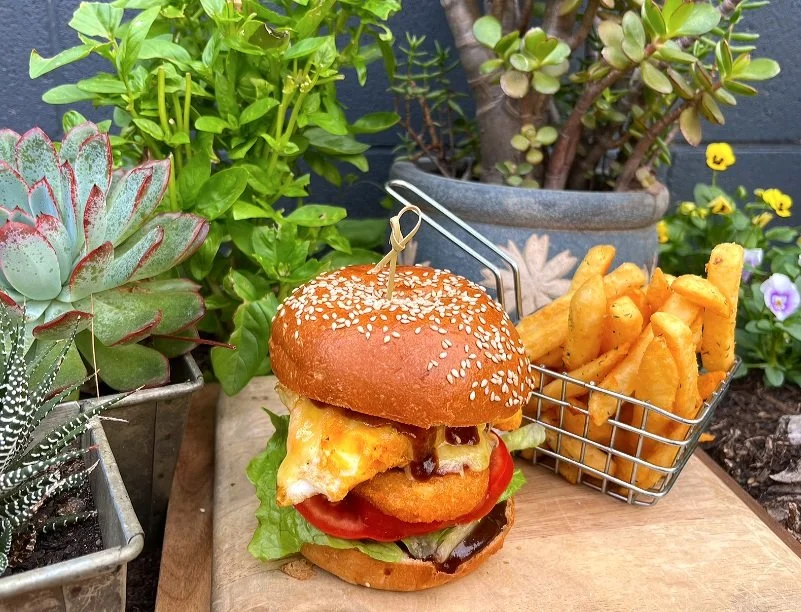 Gourmet burger with lettuce, tomato, fried egg, cheese, and sauce in a sesame seed bun, served with French fries in a metal basket, surrounded by potted plants and flowers.