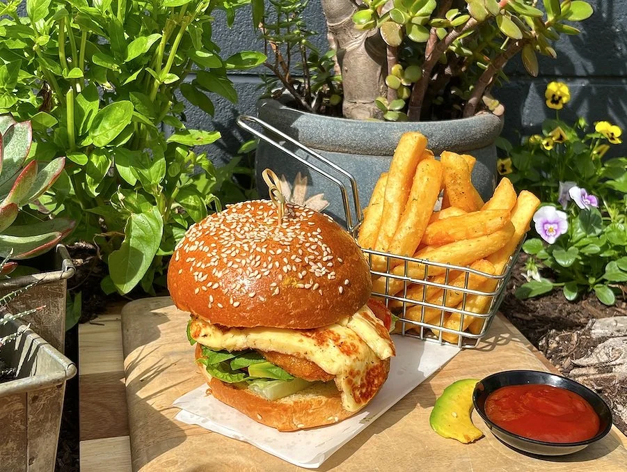A crispy chicken sandwich with lettuce and cheese in a sesame seed bun, served with a side of thick-cut French fries in a metal basket, accompanied by a small bowl of ketchup, set outdoors on a wooden surface surrounded by green plants and flowers.