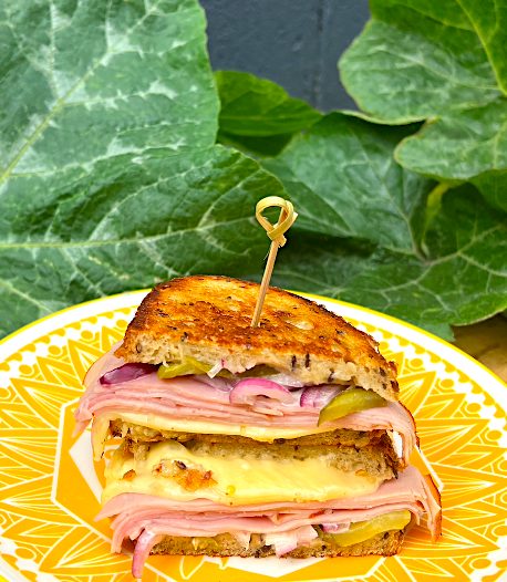 A ham and cheese sandwich with pickles on a yellow and white plate, held together with a toothpick, in front of green leafy plants.