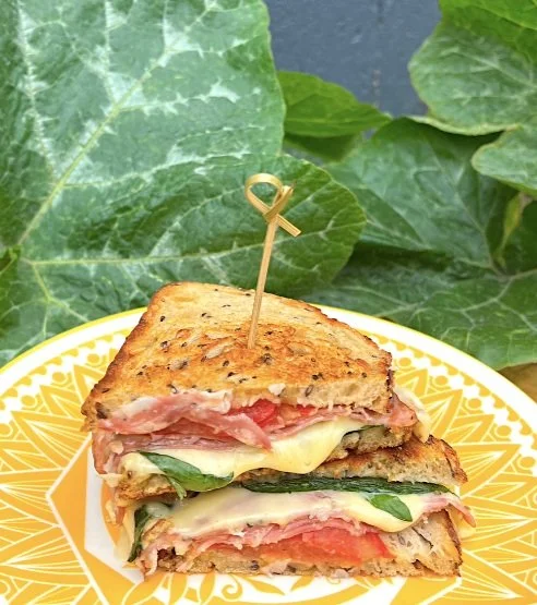 Half of a grilled ham and cheese sandwich with tomato and spinach on a yellow plate, set against green leafy background.