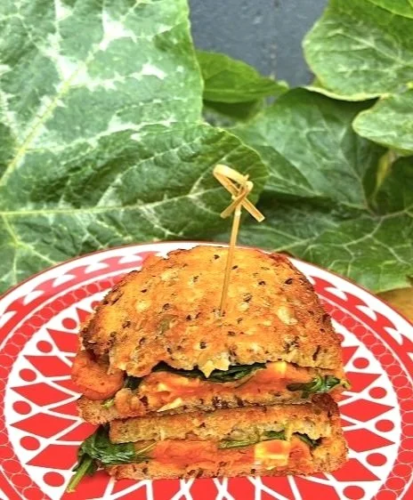 Vegetable sandwich with green leafy vegetables between toasted bread, secured with a toothpick, on a red and white patterned plate, with green leaves in the background.