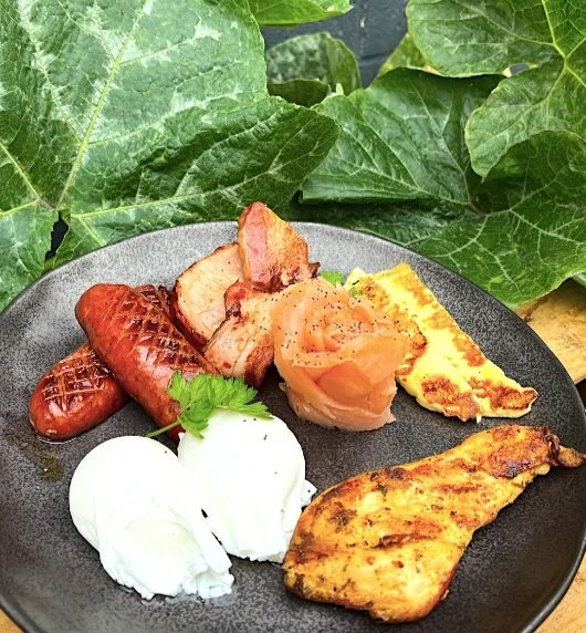 Breakfast plate with sausage, bacon, scrambled eggs, grilled chicken breast, baked potato, and two poached eggs, on a dark plate with green leafy background.