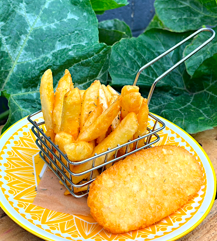 A metal basket of crispy French fries on a plate with a large, golden breaded cutlet beside it. The plate has a yellow and white decorative pattern, set on a wooden surface with green leafy plants in the background.