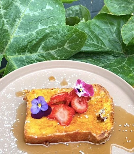 A slice of French toast topped with strawberries and edible flowers, with syrup drizzled on top, on a beige plate surrounded by large green leaves.