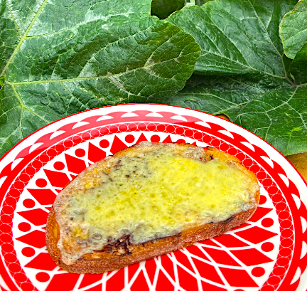 A slice of bread with melted cheese on top, placed on a red and white patterned plate, with green leafy vegetables in the background.