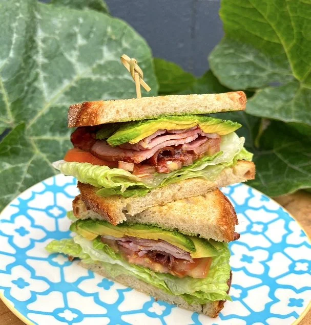 Double-layer sandwich with bacon, lettuce, tomato, avocado, and bread slices on a decorative blue and white plate, set against green leaves in the background.