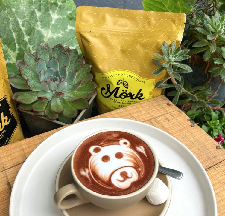 A cup of hot chocolate with latte art of a bear's face, placed on a white saucer with a spoon and a small marshmallow, on a wooden table, with green plants and a yellow packet of Moch chocolate in the background.