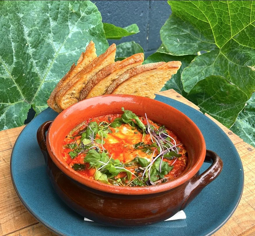 Baked eggs in a tomato sauce garnished with microgreens, served in a brown ceramic bowl with toasted bread slices on the side, placed on a blue plate surrounded by green ivy leaves.