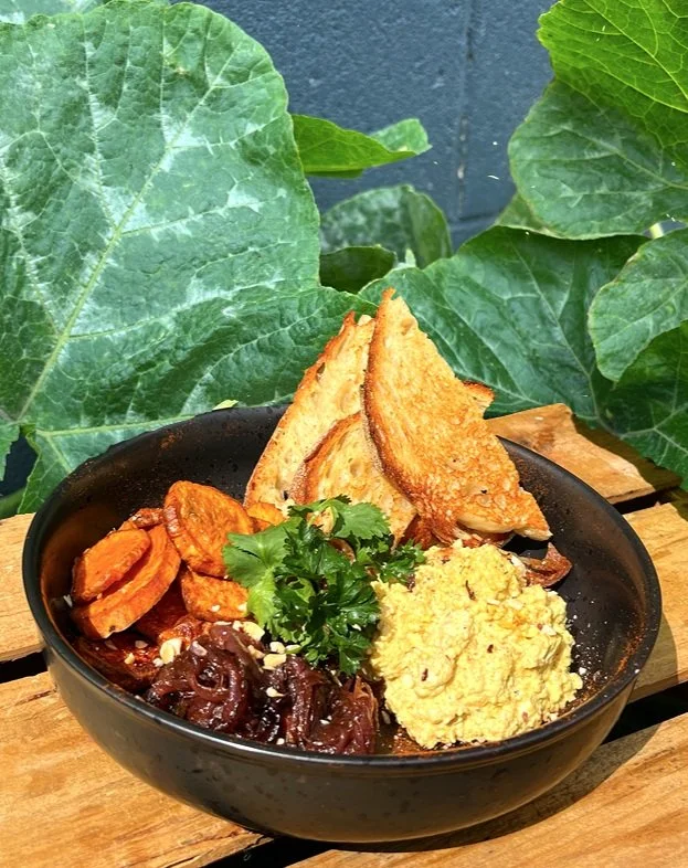 A black bowl of food with toasted bread wedges, plantains, seasoned beans, and a scoop of hummus garnished with cilantro, placed on a wooden surface with green leafy plants in the background.
