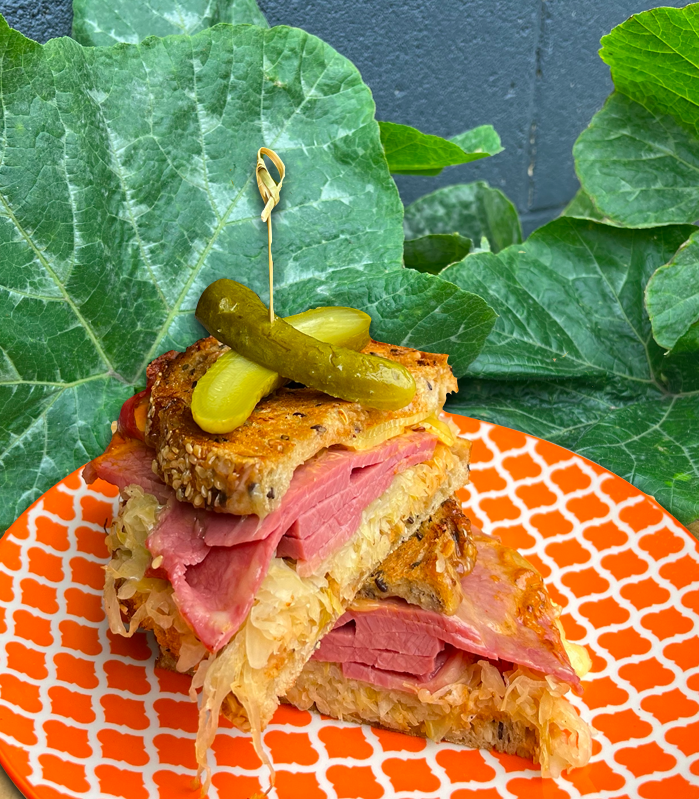 A plate with two pieces of Reuben sandwich topped with pickles and a toothpick, set against a backdrop of green leaves.