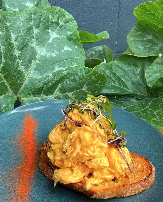 Open-faced toast topped with scrambled eggs and microgreens, served on a blue plate with a sprinkle of red spice, with large green leaves in the background.