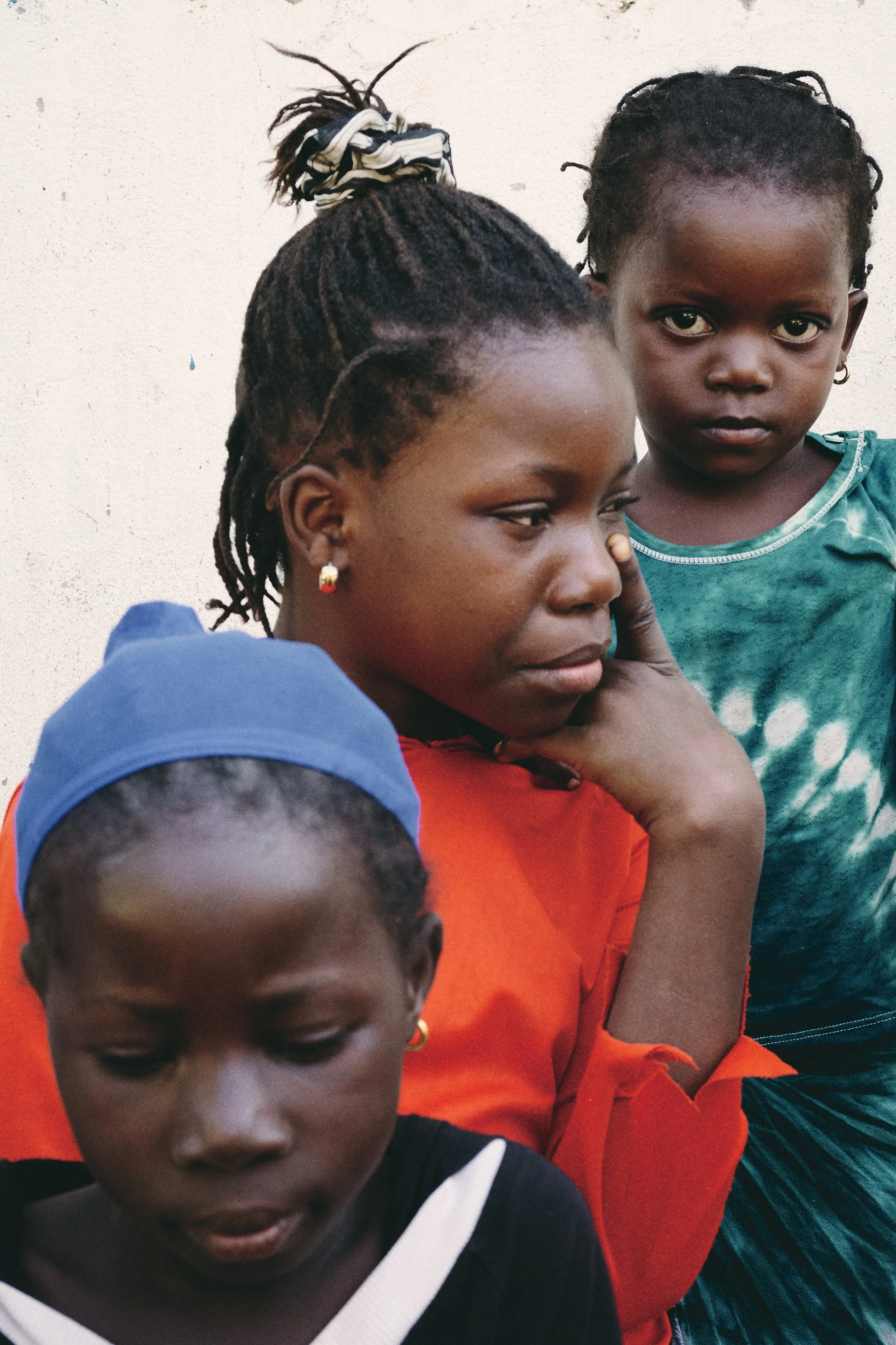 Three young girls with dark skin and braided hair standing close together against a plain white wall. One girl is wearing a teal tie-dye shirt, another a bright orange shirt, and the third a black shirt with white trim and a blue head covering. The g