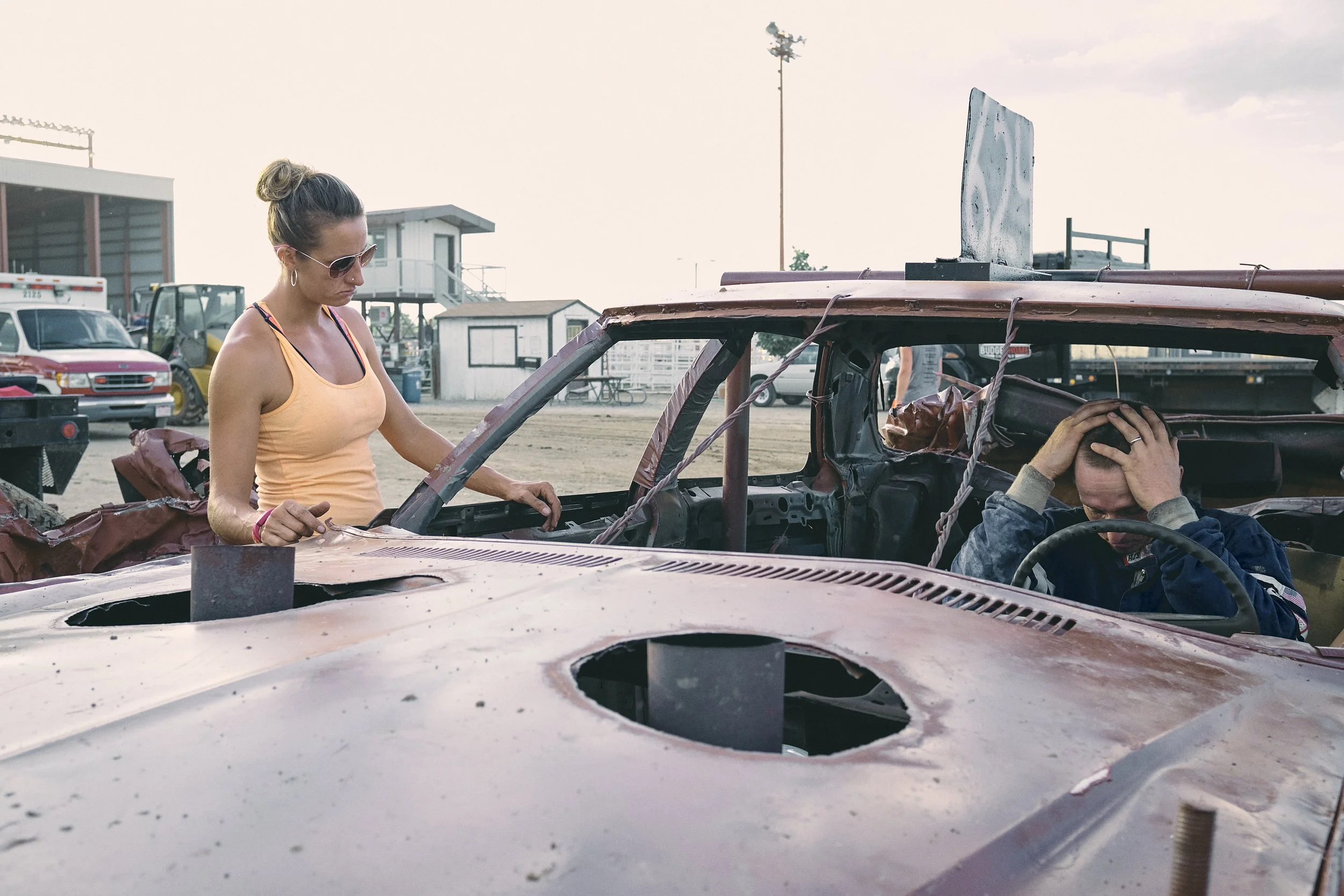 A woman in a tank top and sunglasses looks at a wrecked, demolition derby car with a man inside holding his head in frustration at Boulder County fairgrounds.