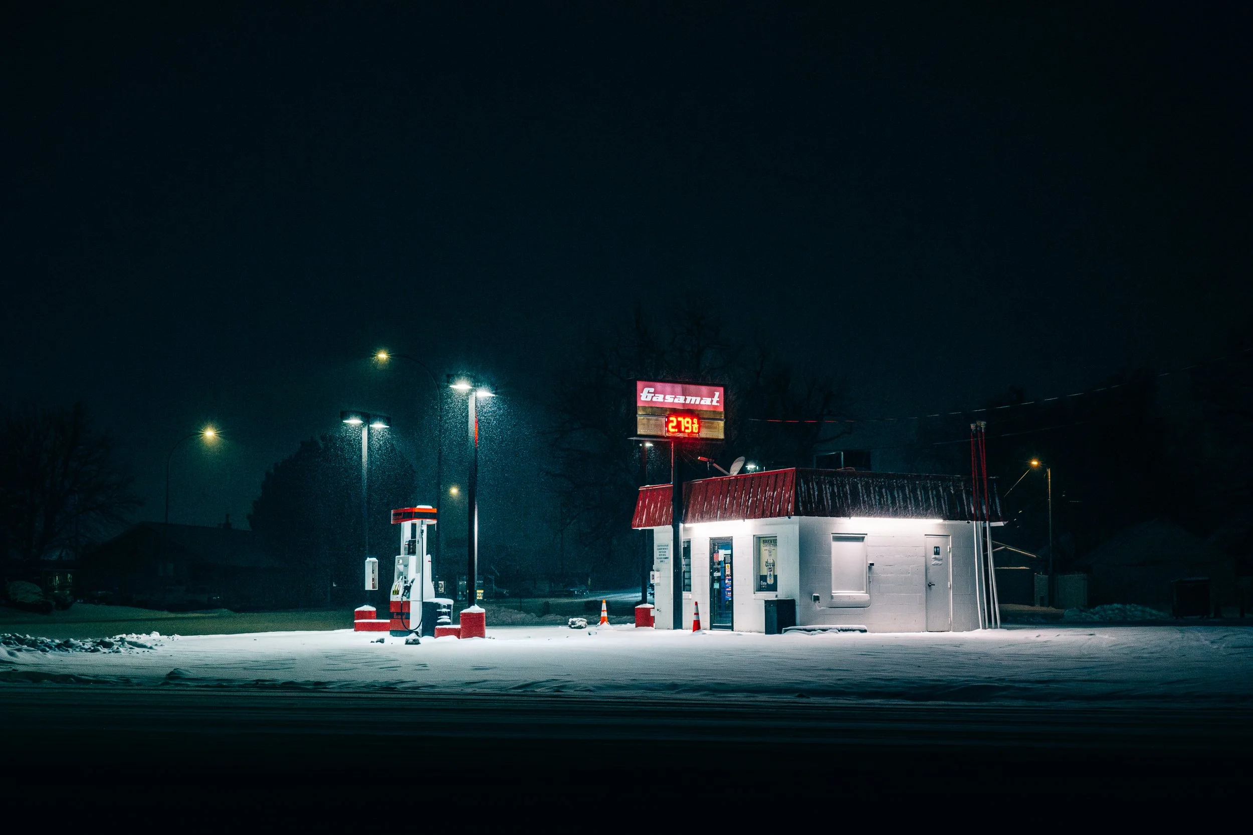 A snow-covered gas station in Longmont Colorado at night illuminated by lights, showing fuel pumps, a small building, and a digital price sign displaying $2.79 per gallon.