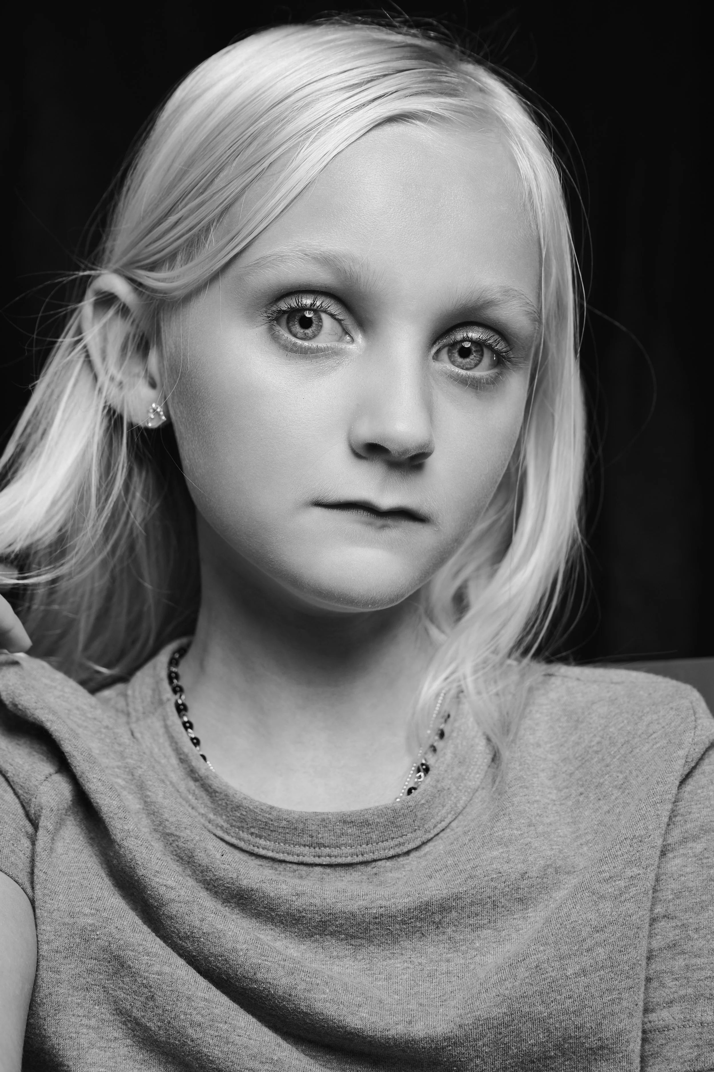 A black and white close-up portrait of a young girl with blonde hair, wearing a necklace and earrings, with a serious expression.