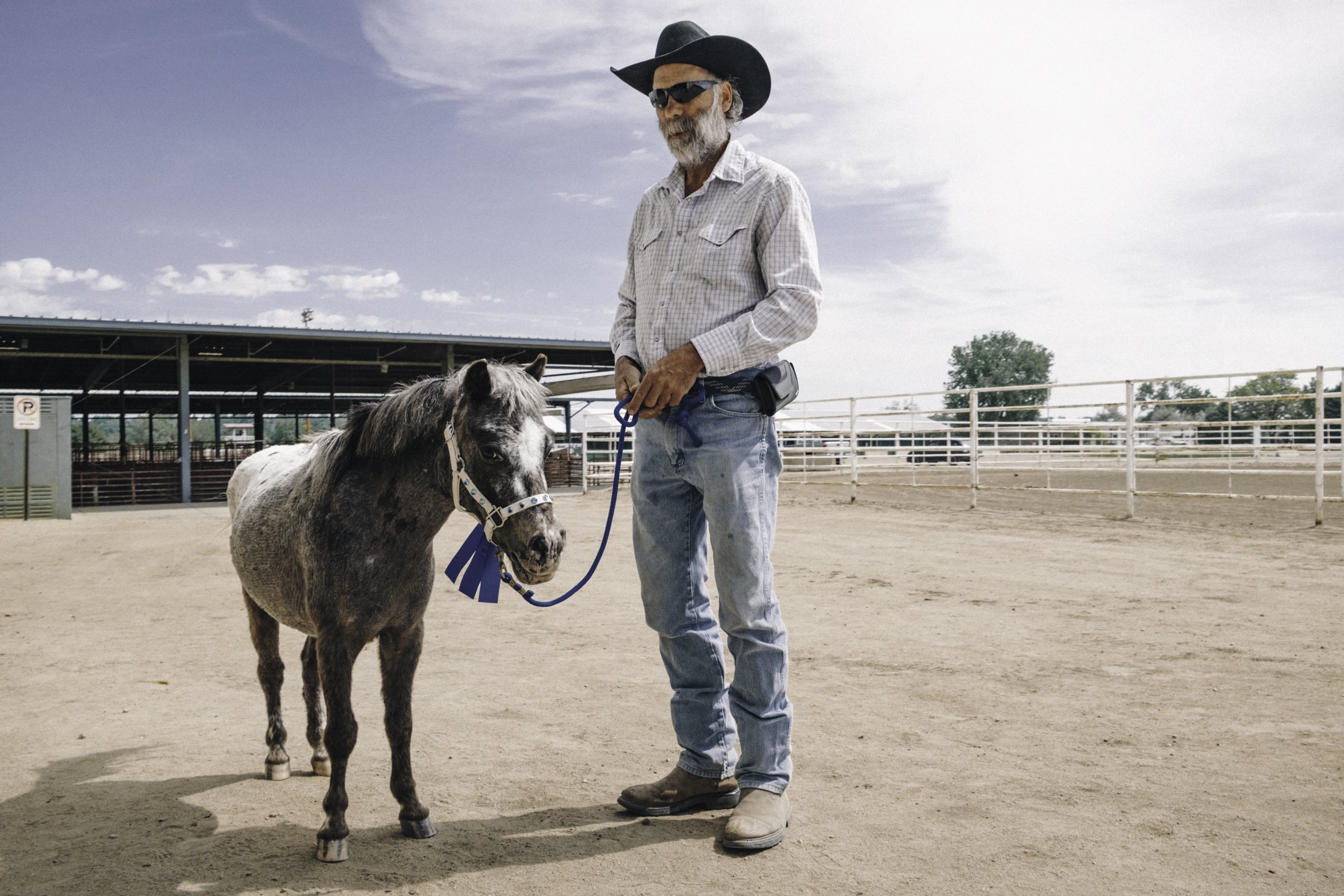 A man wearing a black cowboy hat, sunglasses, a checkered shirt, and jeans stands next to a miniature gray and white horse on a blue lead rope in a sandy outdoor arena.